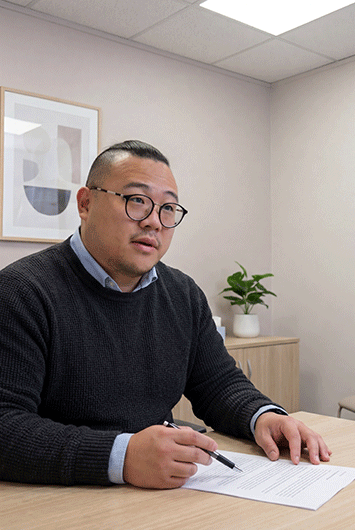 Consultant surgeon Mr Chad Chang, wearing glasses and a dark jumper over a collared shirt, sitting at a desk and reviewing a clinical document with a patient during a consultation.