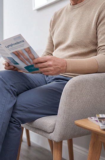 A patient wearing a medical-grade compression sleeve on their left forearm while reading a clinical educational brochure titled "Lymphoedema" in a professional waiting area.