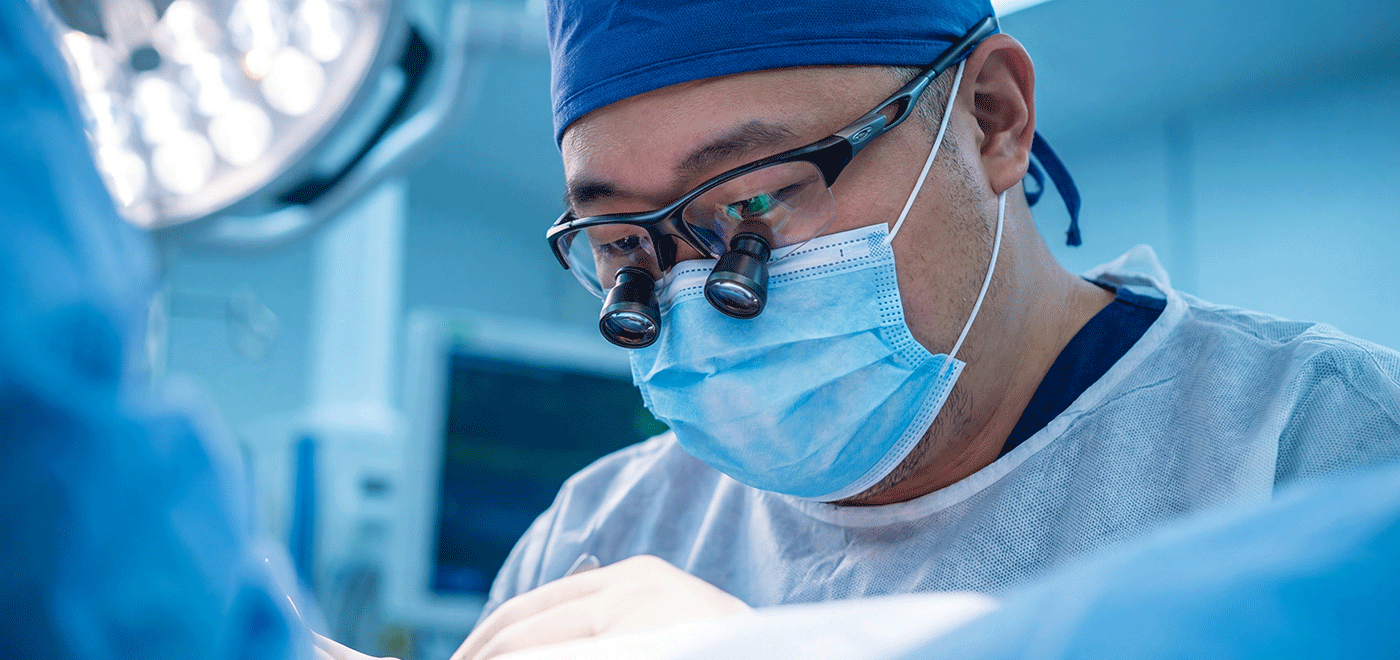 A close-up of consultant surgeon Mr Chad Chang wearing surgical loupes, a mask, and a blue scrub cap, performing a precision lymphatic debulking procedure under high magnification.