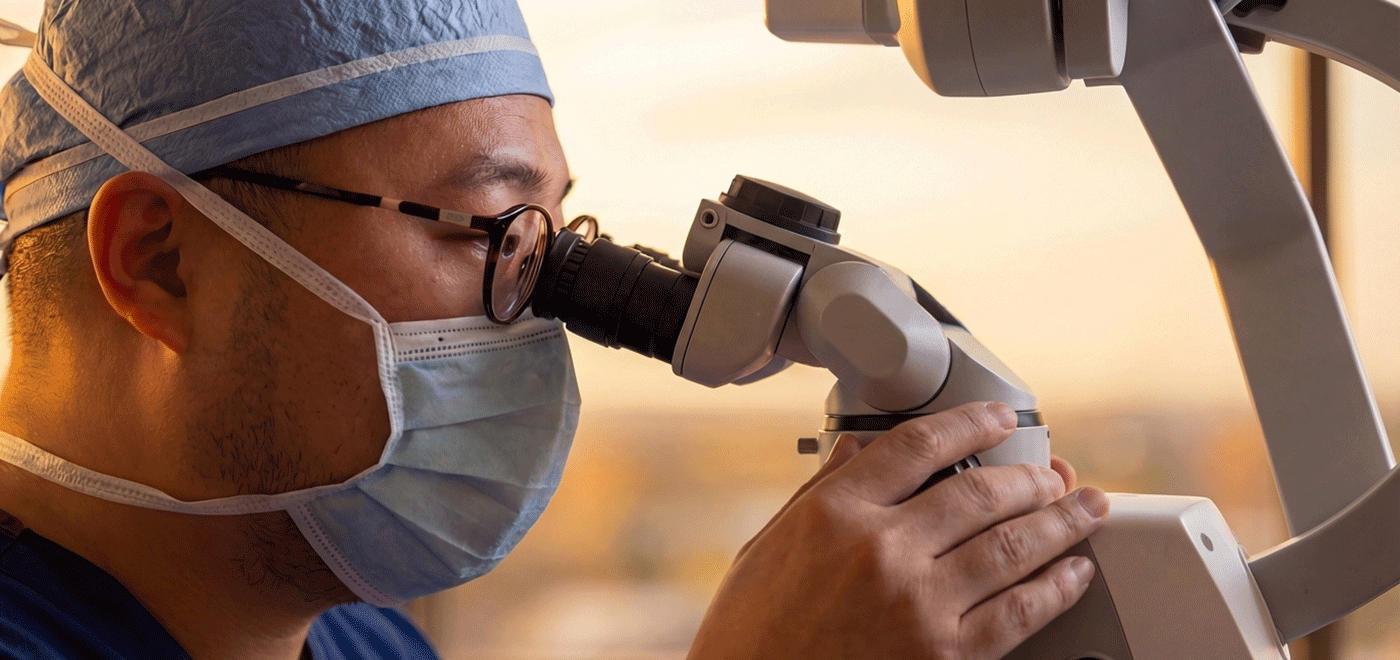 A close-up profile of consultant surgeon Mr Chad Chang wearing a surgical mask and glasses, performing a pre-operative setup and calibration of a high-magnification operating microscope for a supermicrosurgical Lymphaticovenous Anastomosis (LVA) procedure.